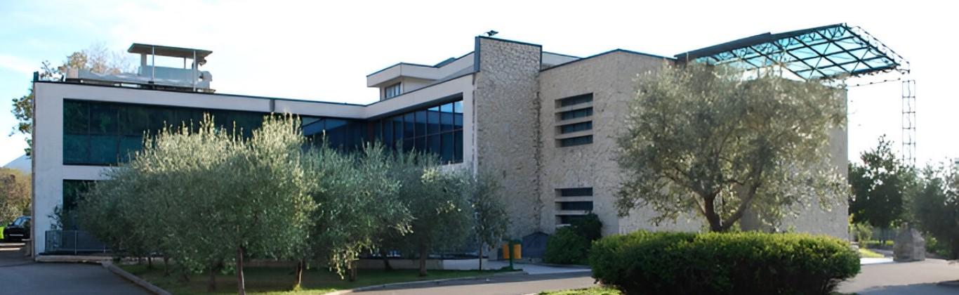 Edificio moderno con ampie vetrate, circondato da alberi e vegetazione.